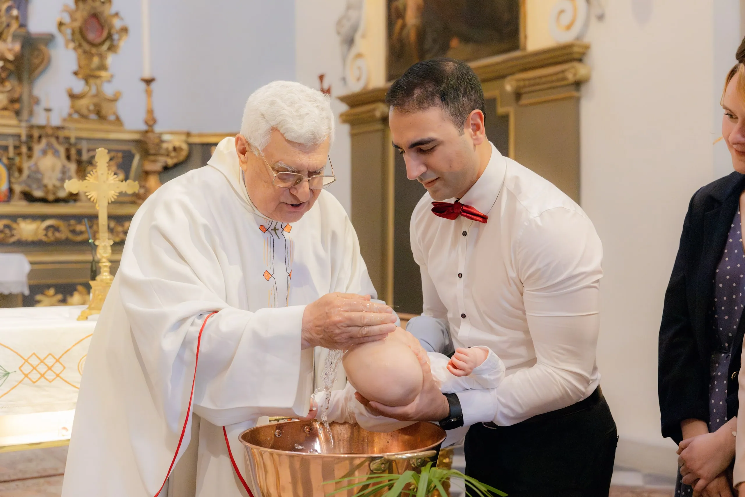 Le prêtre verse l'eau du baptême sur le front du bébé, que son père tient au-dessus de la vasque en cuivre