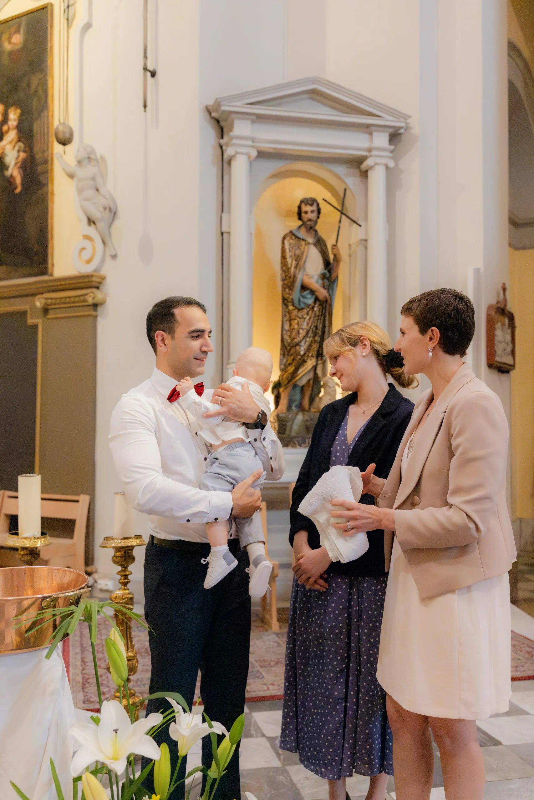Le père au nœud papillon rouge porte son bébé, face à deux femmes dont l'une tient un linge blanc