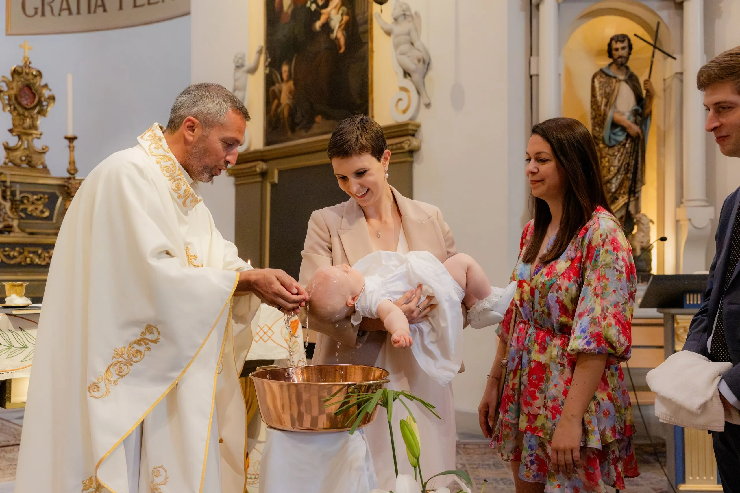 Le prêtre en chasuble blanche brodée d'or verse l'eau sur la tête d'un bébé que porte une femme en veste beige, une femme en robe fleurie à côté
