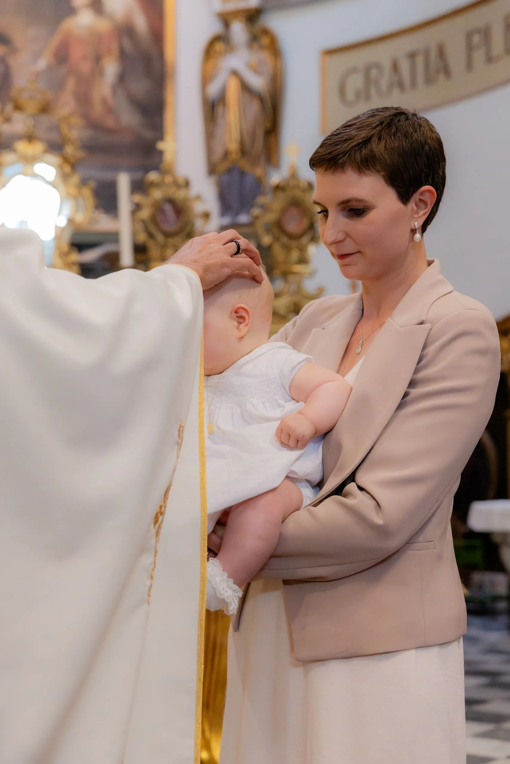 Le prêtre pose la main sur la tête du bébé, tenu dans les bras d'une femme en veste beige
