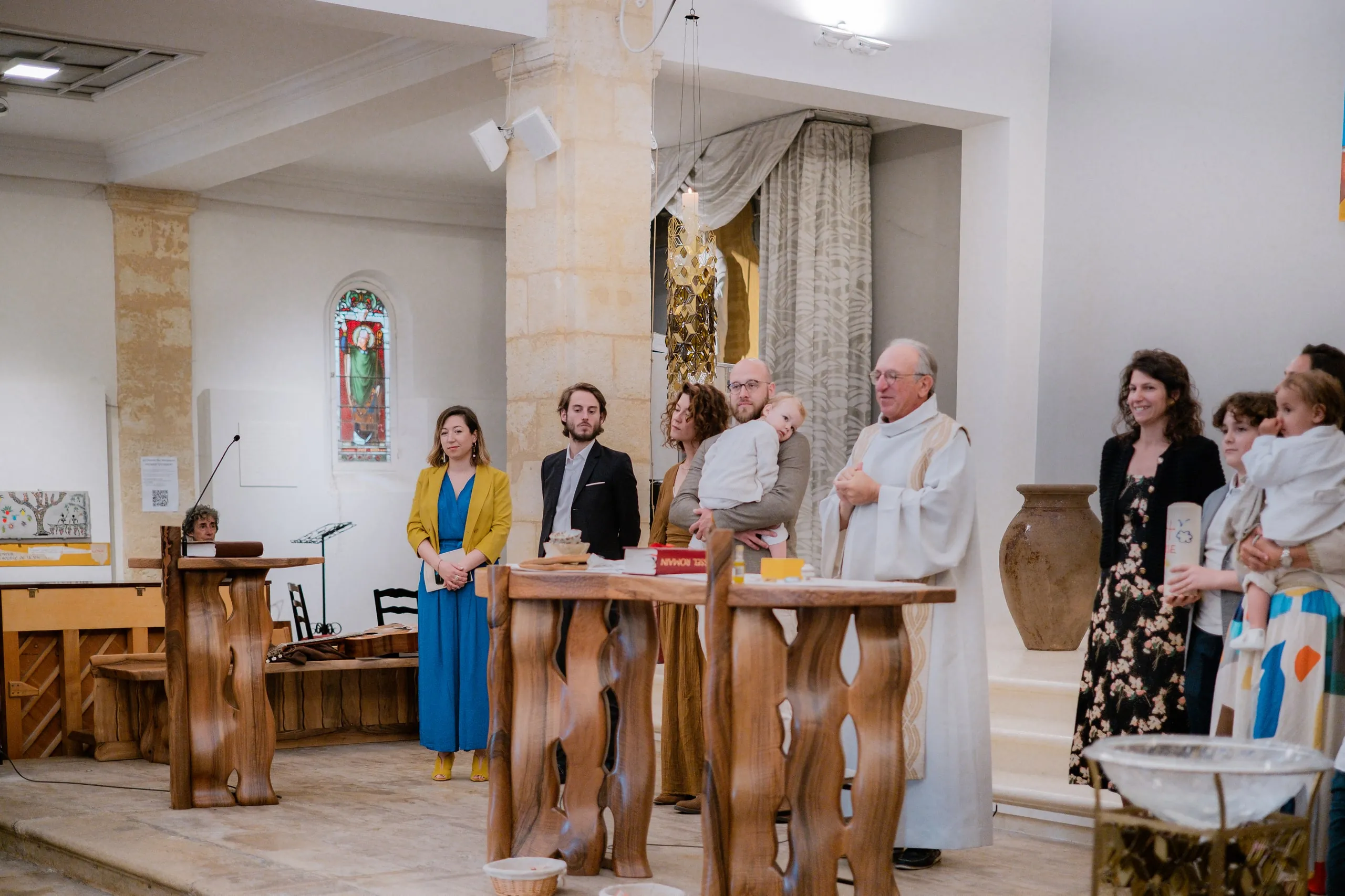 Dans la chapelle, le prêtre en aube blanche derrière l'autel en bois, entouré de la famille et des proches, un père portant son petit garçon