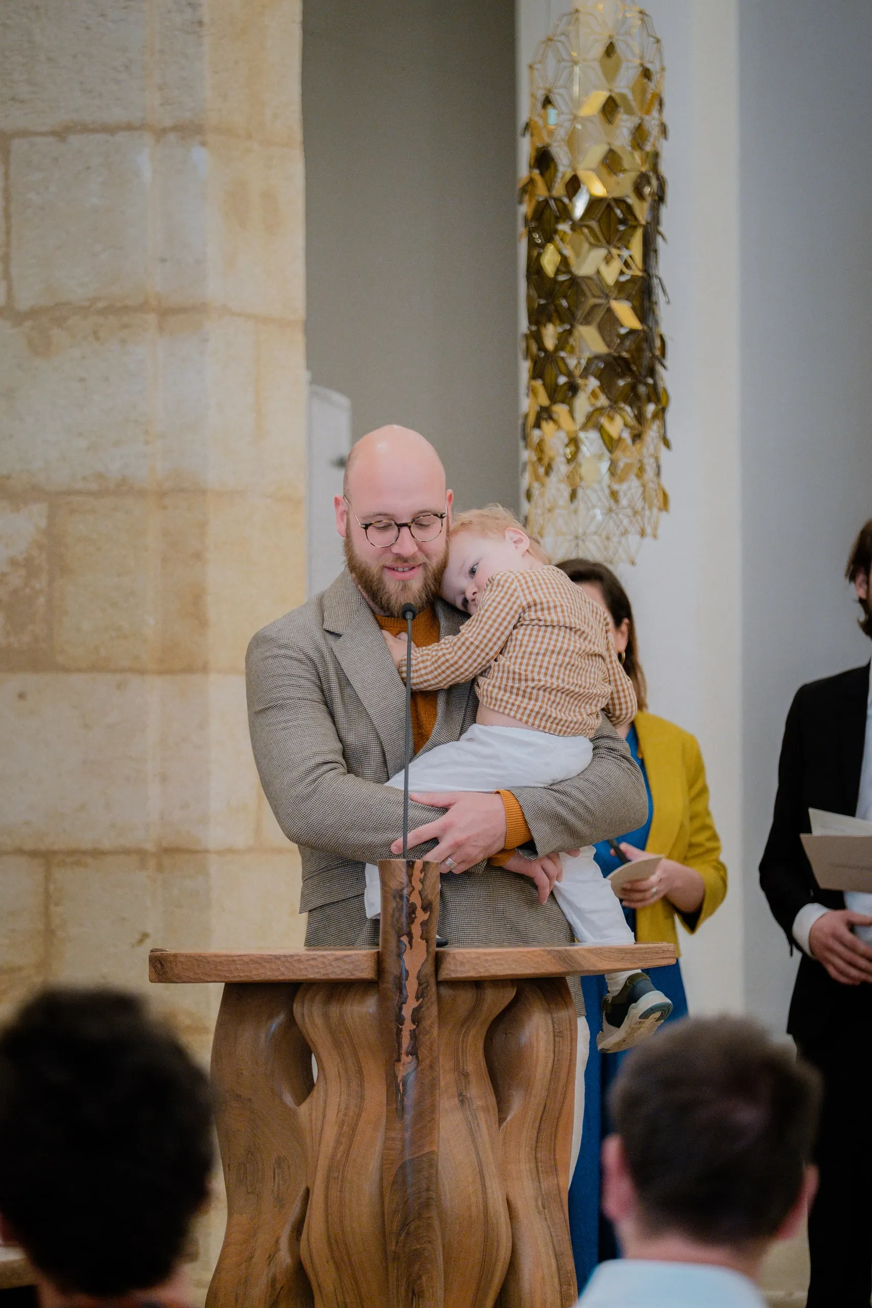 Le père tient son petit garçon contre lui derrière le pupitre en bois, entouré de proches