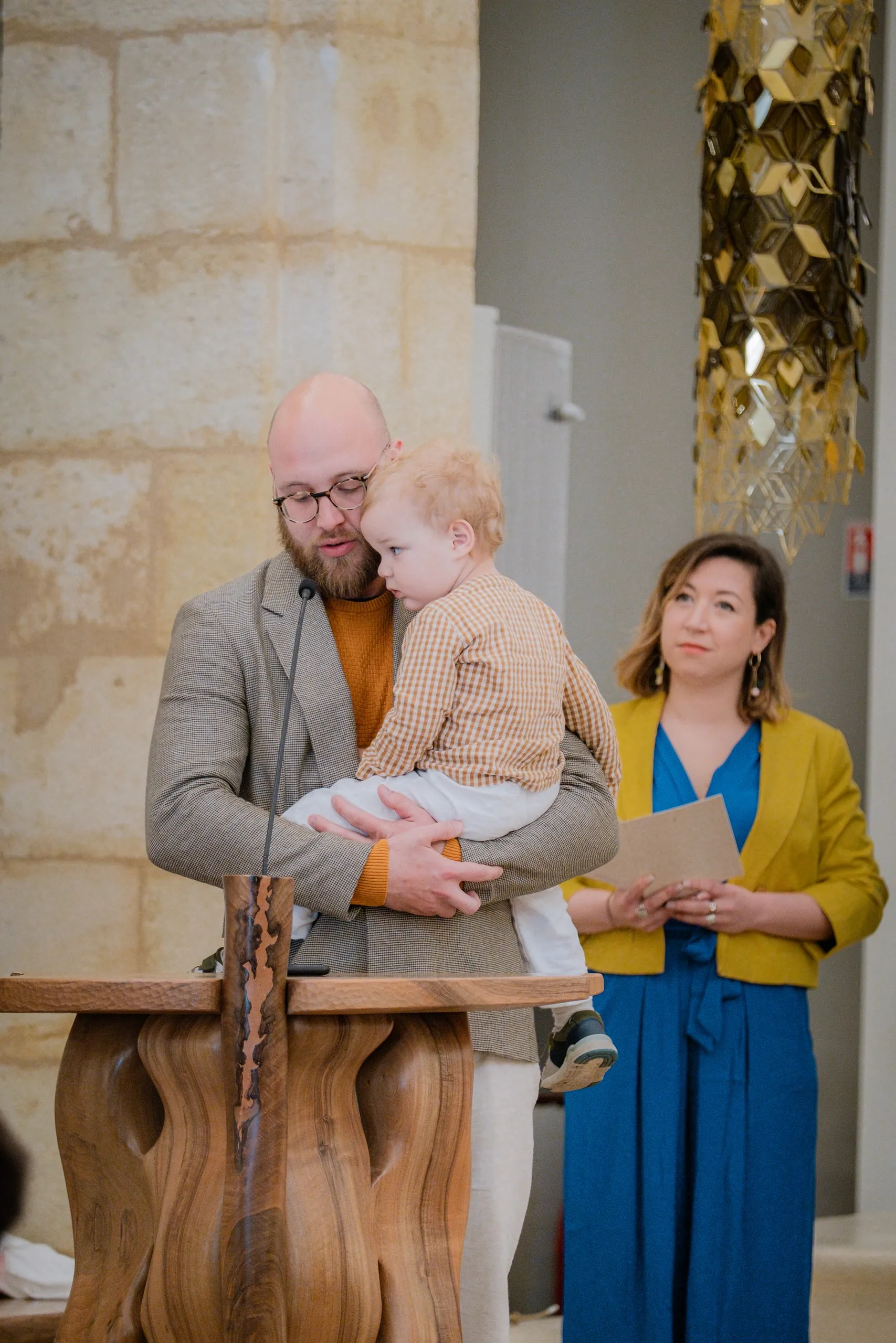 Le père porte son petit garçon derrière le pupitre, à côté d'une femme en veste jaune