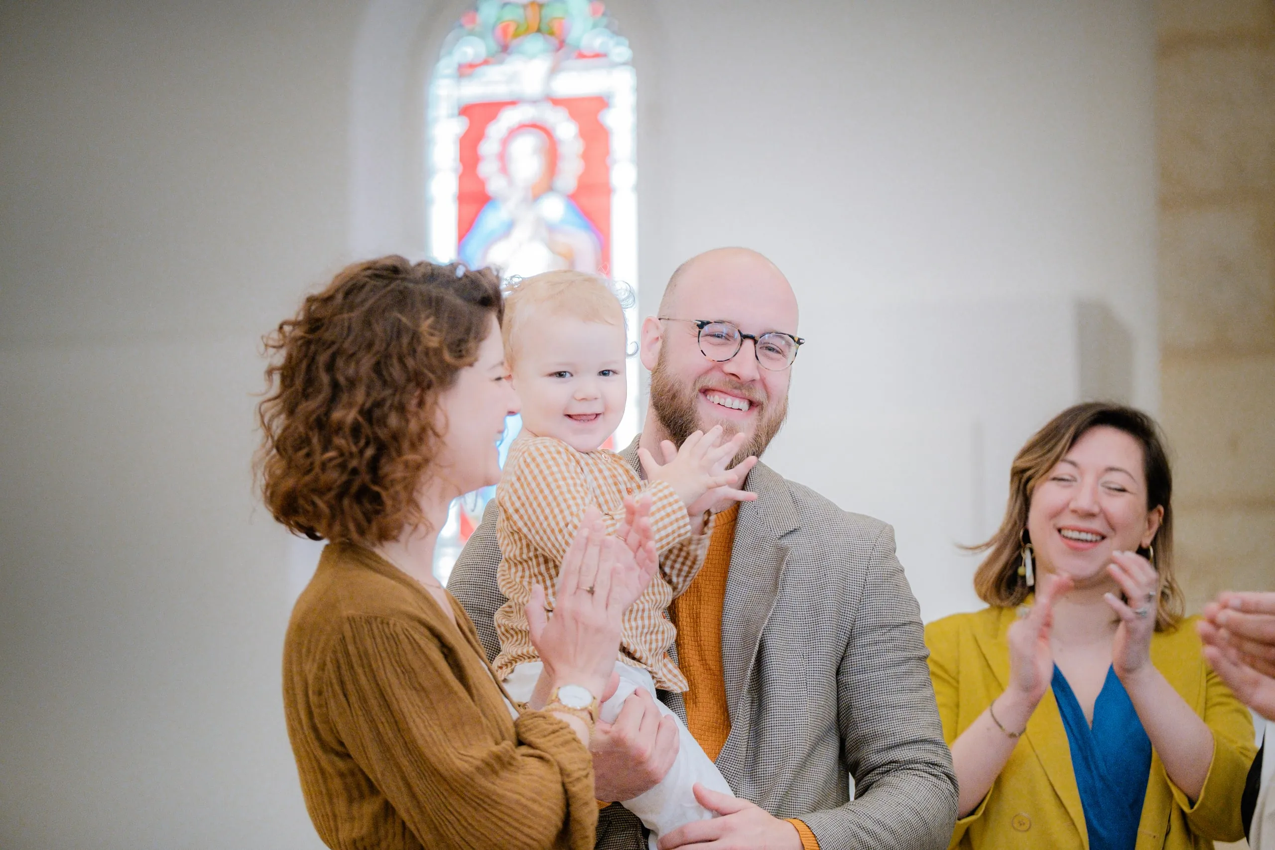 Le petit garçon rit aux éclats en applaudissant, entouré de ses parents et d'une femme en veste jaune qui rit et applaudit, devant un vitrail