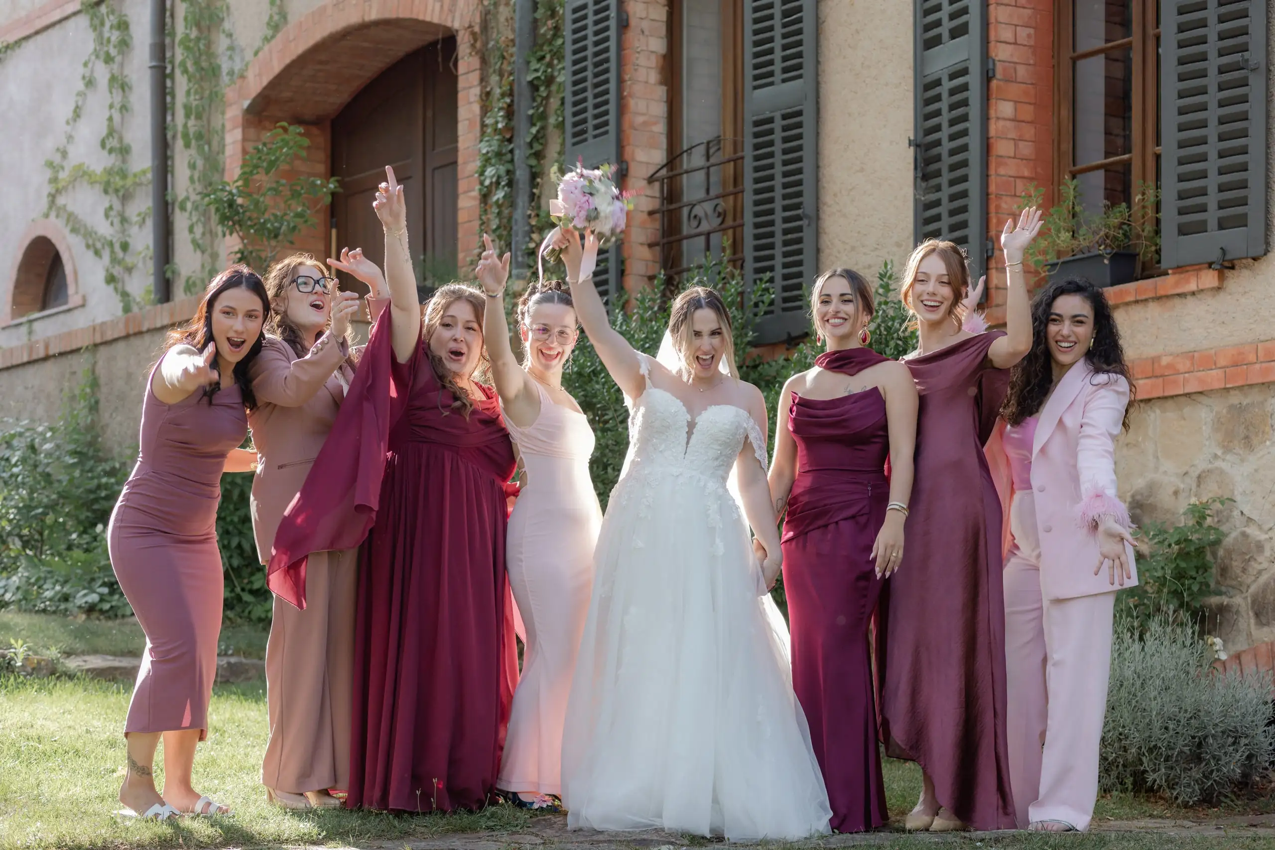 La mariée et ses demoiselles d'honneur en robes roses et prune, bras levés devant la bastide