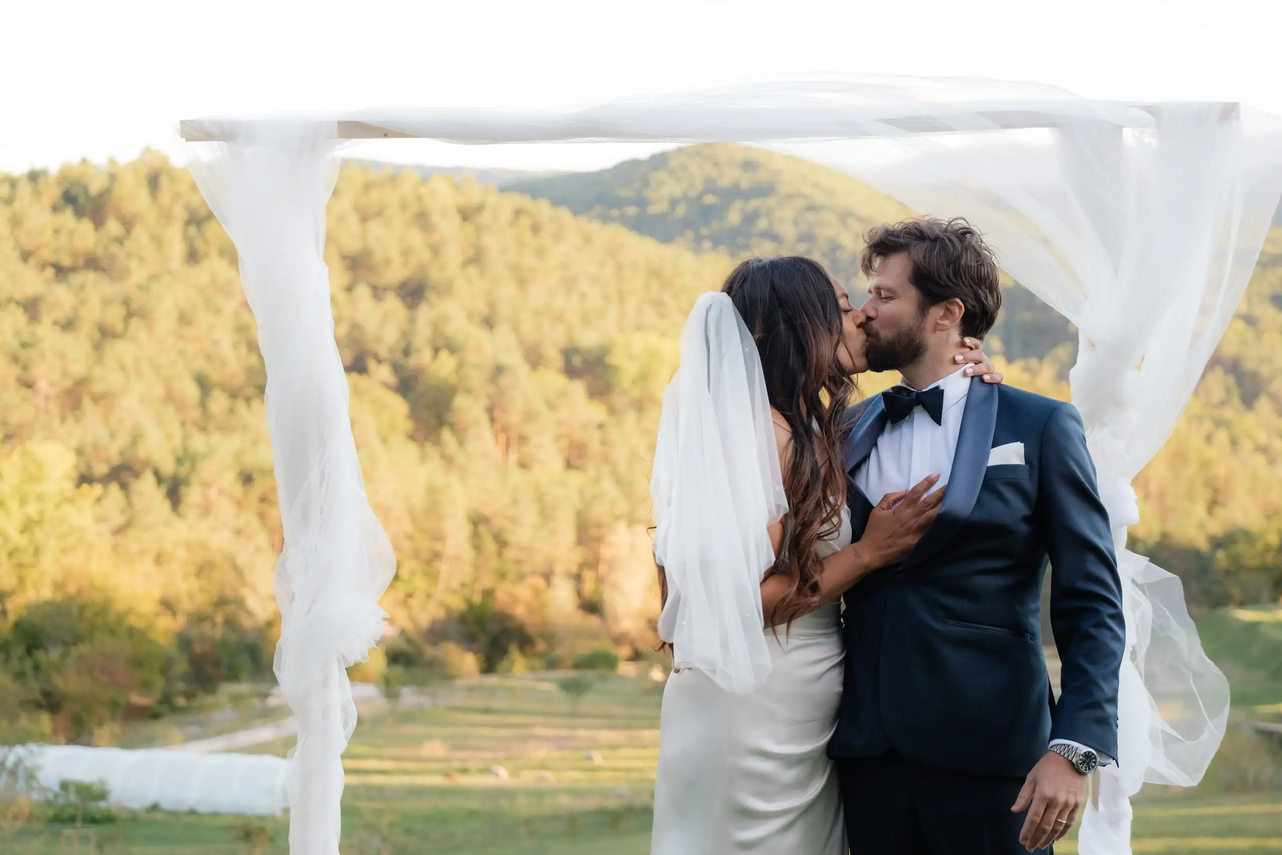 Les mariés s'embrassent sous une arche de voiles blancs, les collines derrière eux